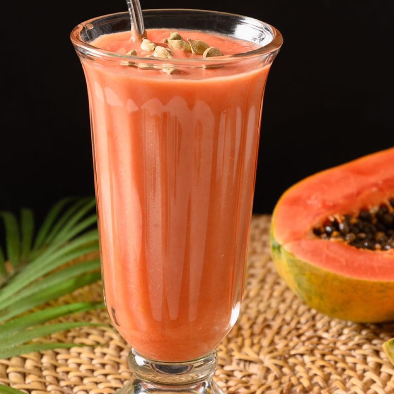 Papaya smoothie in a glass with cardamom pods, next to a halved papaya and palm leaf.