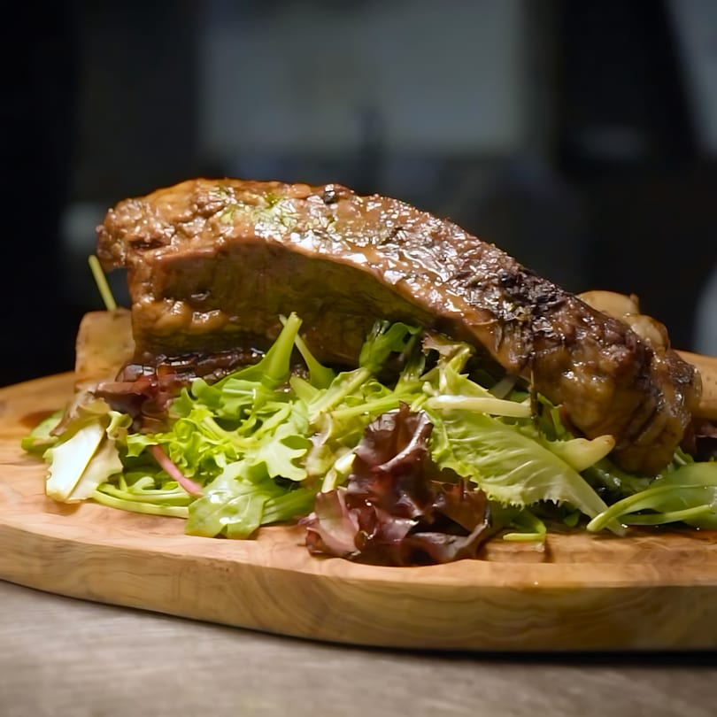 Braised short rib with a side of fresh mixed greens, served on a wooden board.