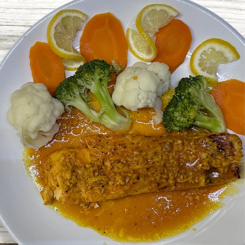 Dominican salmon dish with orange sauce, broccoli, cauliflower, carrots, and lemon slices on a white plate.