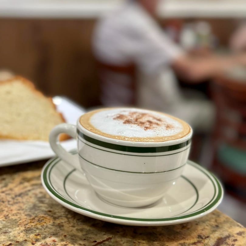 A cappuccino with foam and cinnamon in a white cup with green stripes, next to a slice of cake.