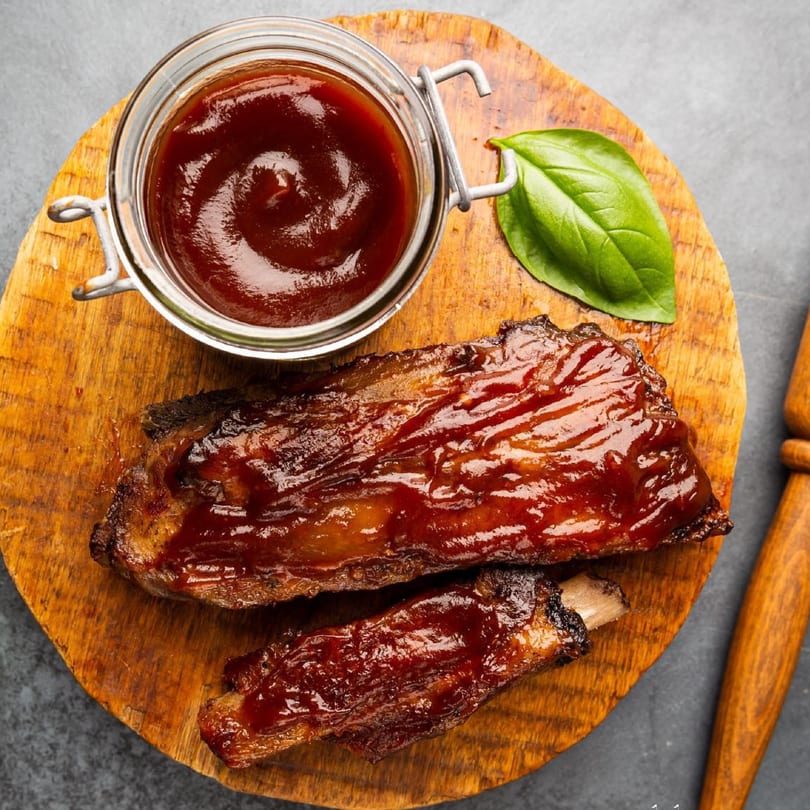 Two glazed ribs and a jar of BBQ sauce on a wooden board with a basil leaf.