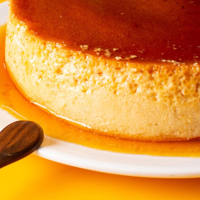Close-up of a golden-brown flan with glistening caramel sauce on a white plate.