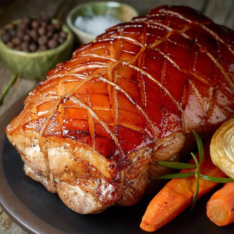 Close-up of a glazed, scored pork roast with carrots, onions, and rosemary on a dark plate.